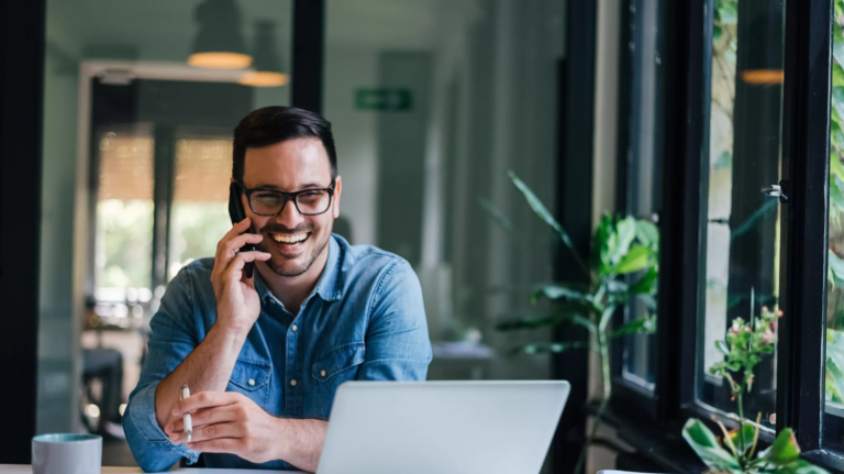 Négociateur immobilier indépendant travaillant sur son site web depuis un bureau, souriant au téléphone devant un ordinateur portable.